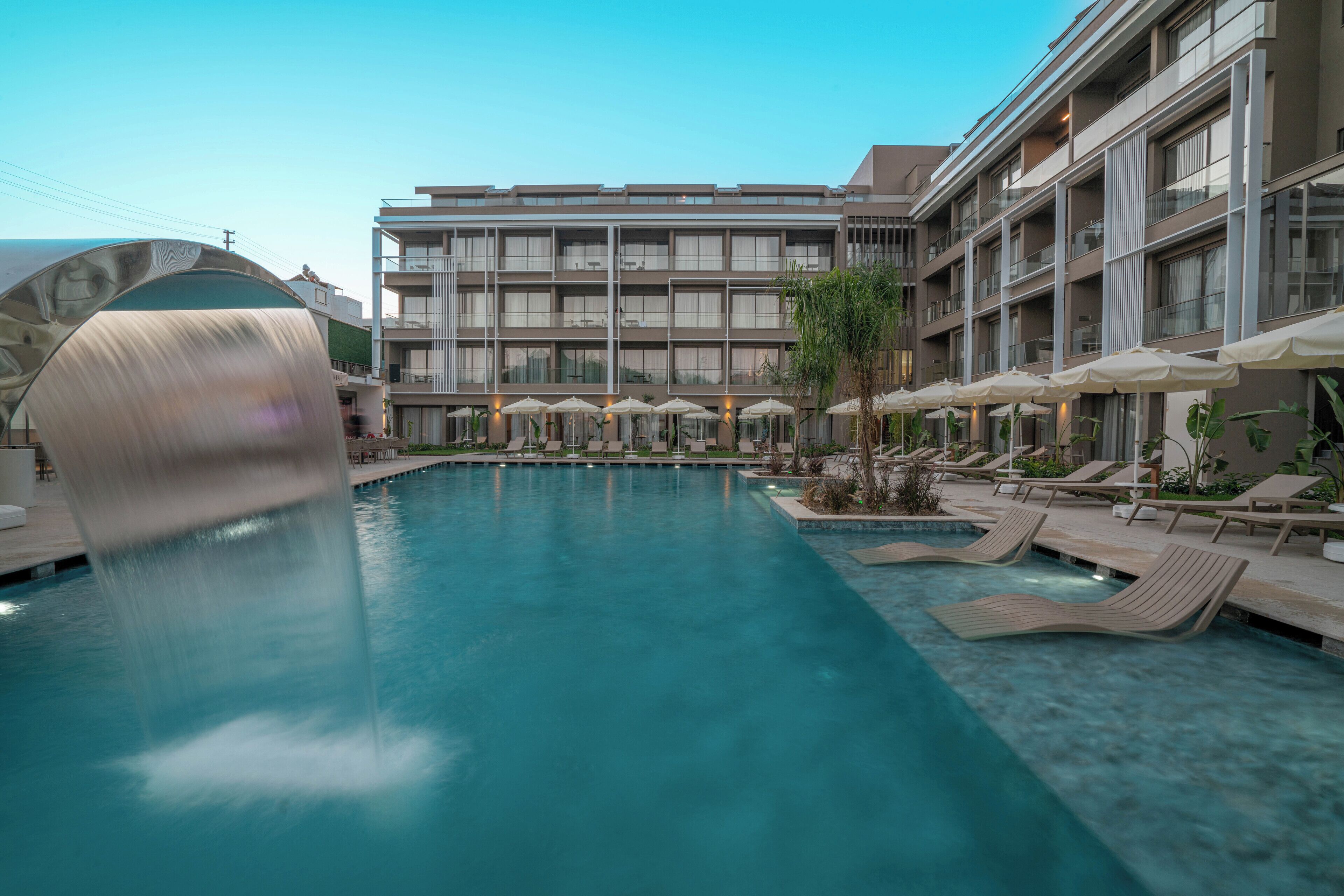 A swimming pool with lounge chairs and umbrellas, surrounded by a multi-story building with balconies.