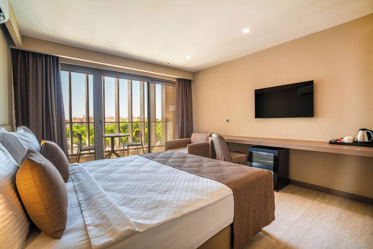 A modern hotel room with a large bed, a desk, a chair, a flat-screen TV, and a balcony with a view of the city.