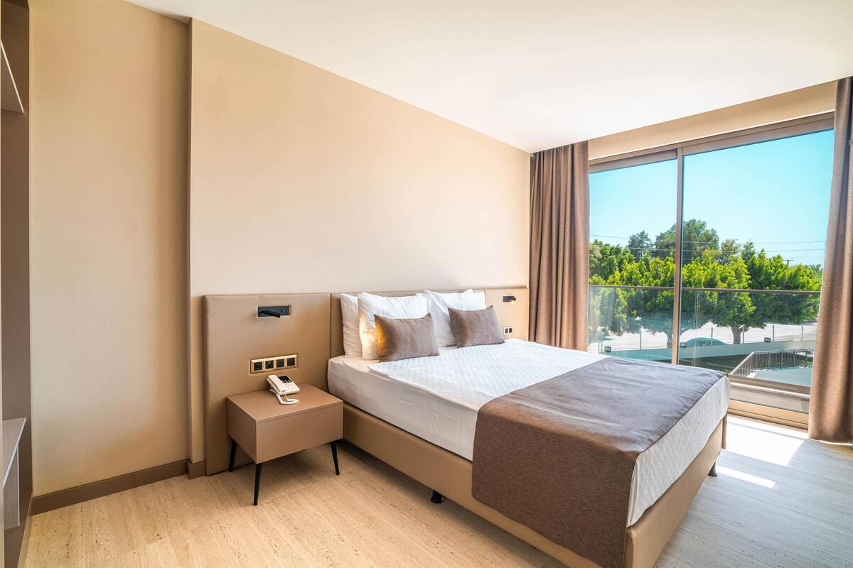 A modern hotel room with a large bed, bedside table, and a view of greenery through the window.