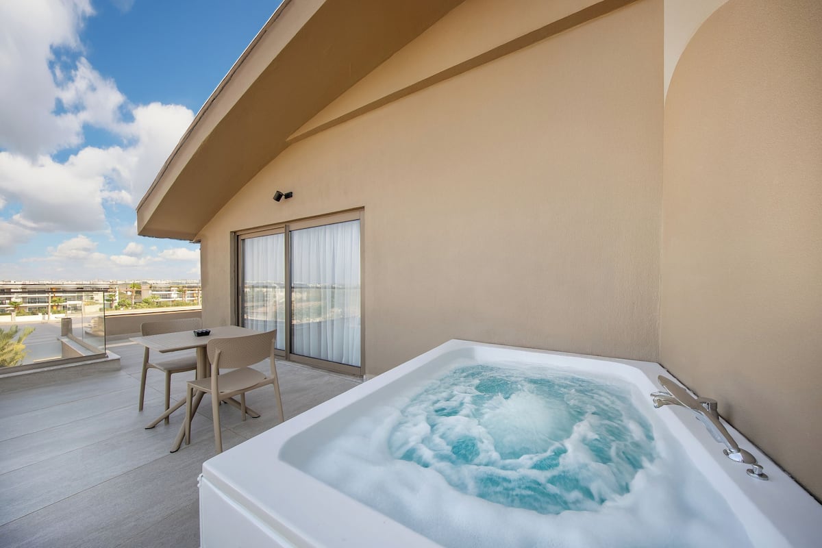 A hot tub on a balcony with a table and chairs, a sliding glass door, and a view of the cityscape.