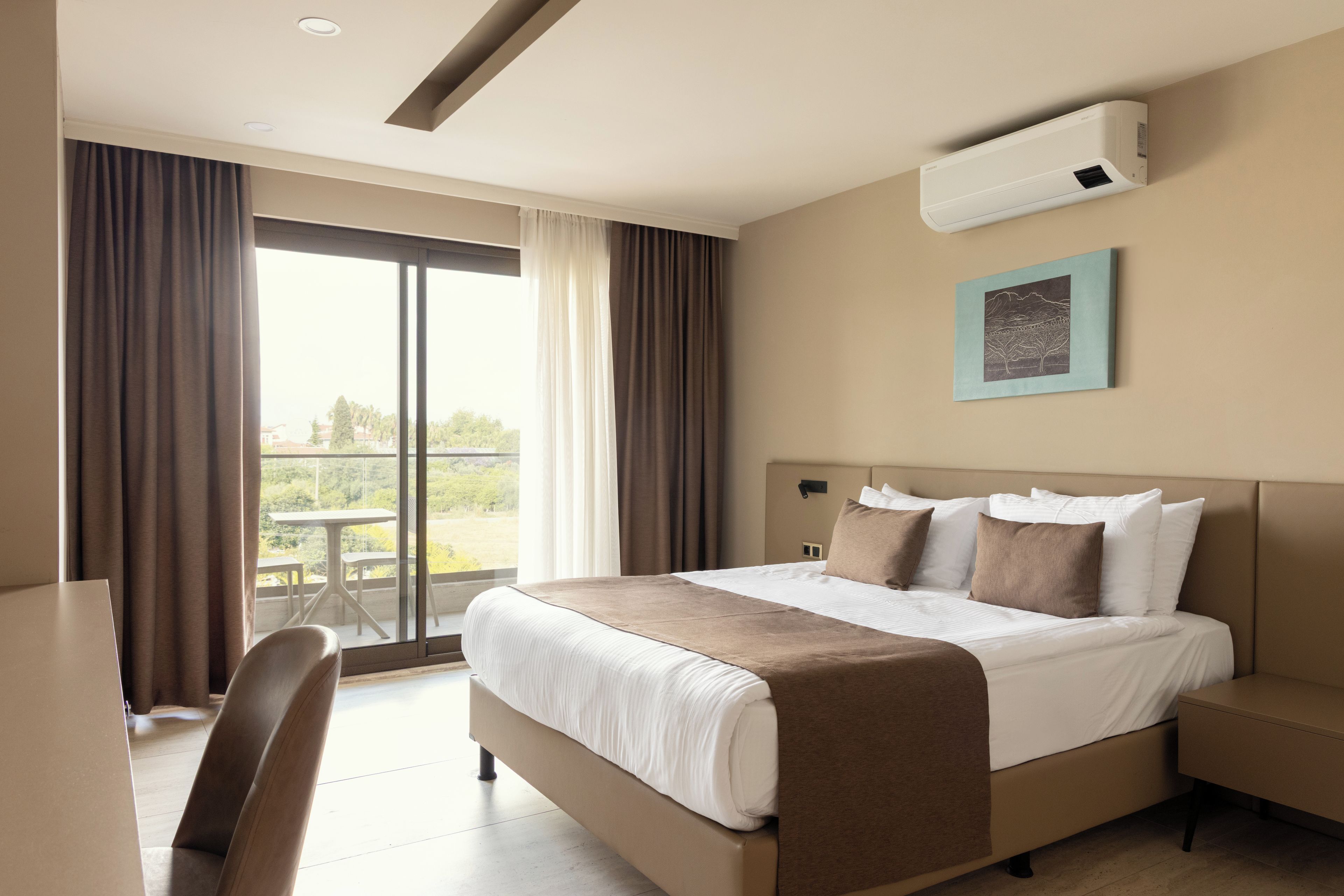 A hotel room with a bed, a desk, and a chair. There is a window with curtains, an air conditioning unit on the wall, and a painting above the bed.