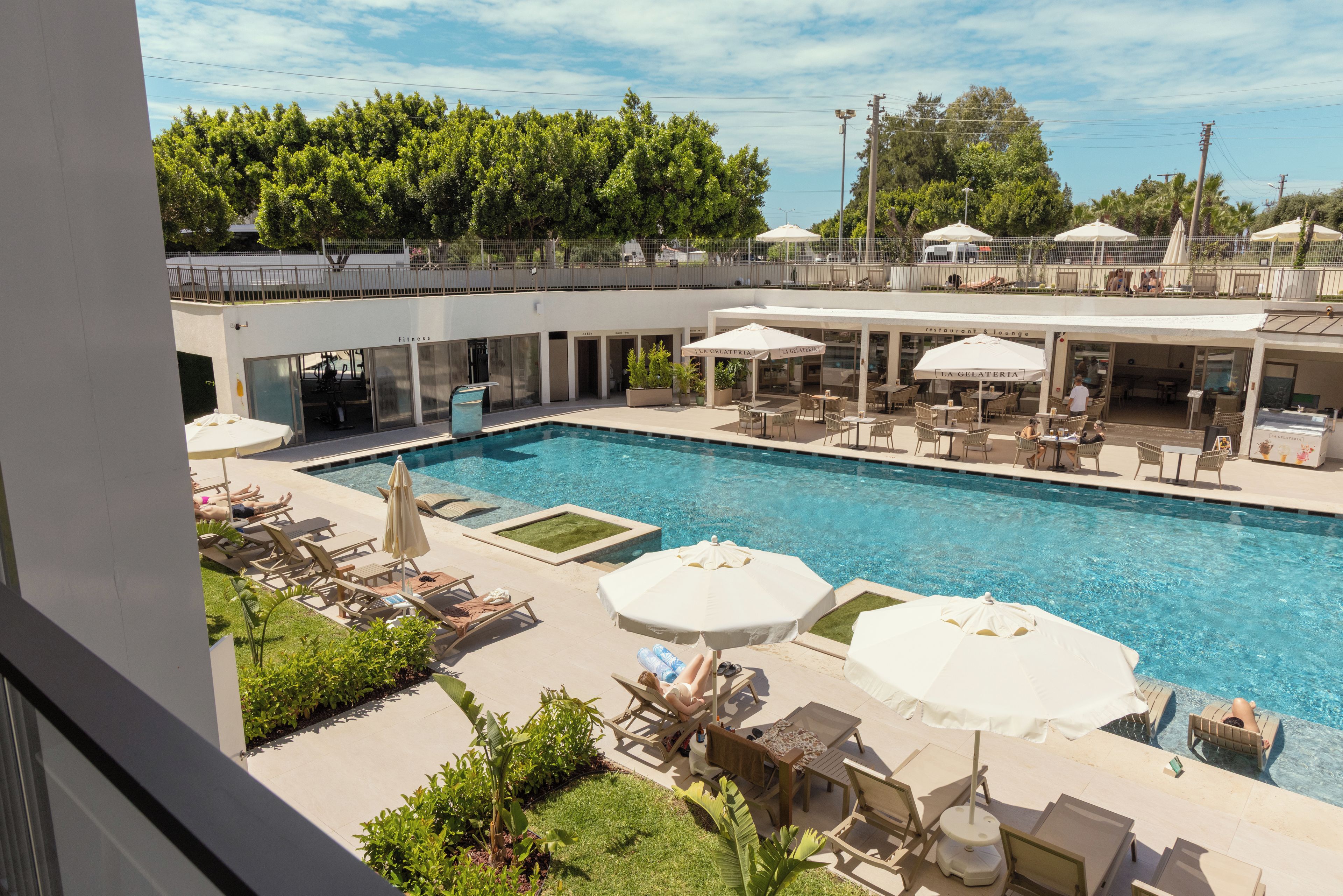 Una piscina con sillones de descanso y sombrillas, rodeada por una zona vallada que incluye un restaurante y un gimnasio.
