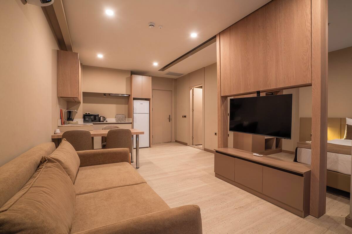 A modern living room with a sofa, a TV mounted on a wooden cabinet, and a kitchen area in the background.