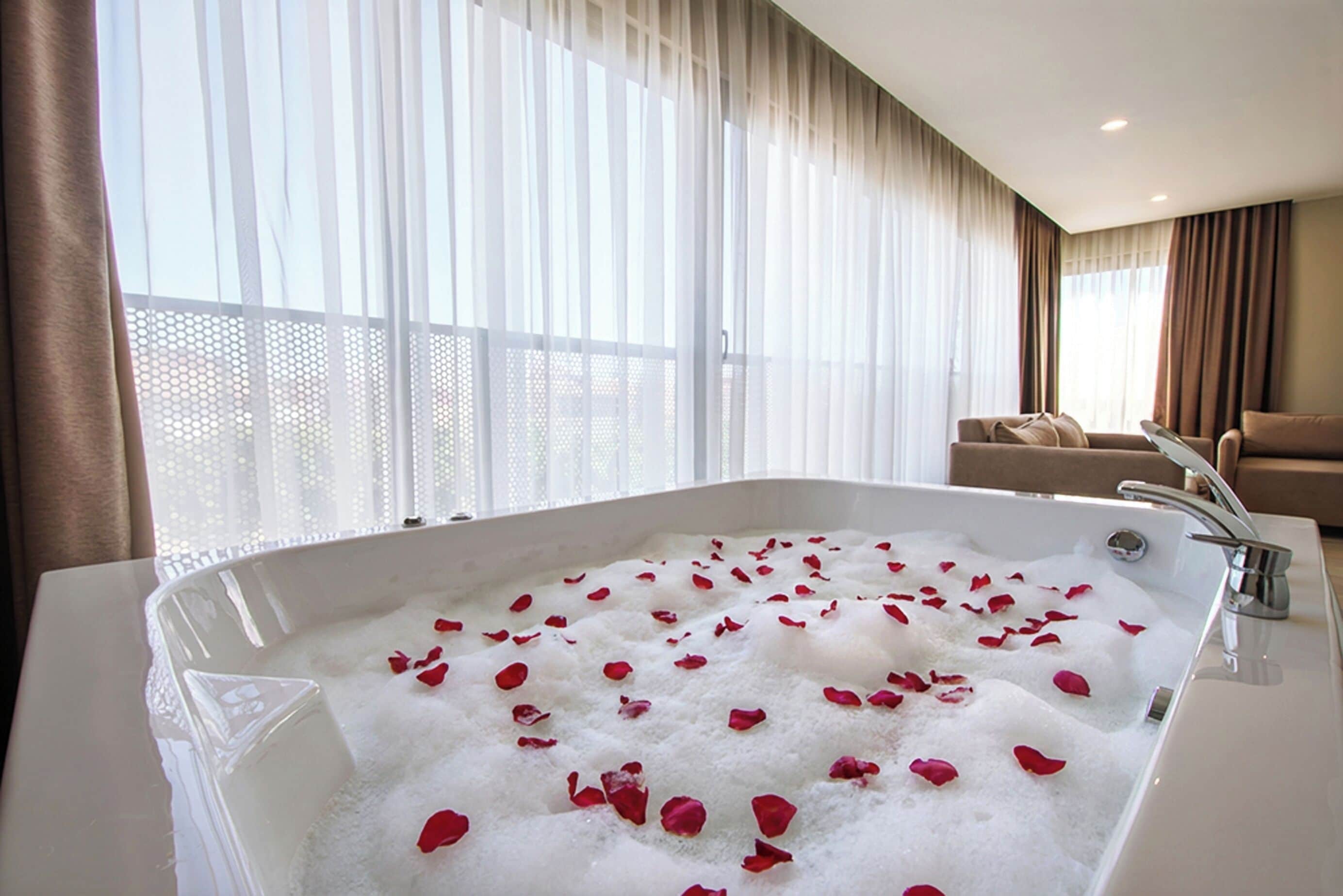 Un baño moderno con una bañera grande llena de burbujas y pétalos de rosa, un sofá y vistas a través de cortinas traslúcidas.