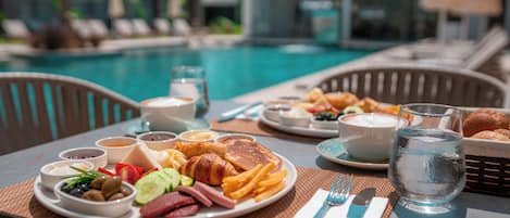 Desayuno junto a la piscina, con platos de comida, un vaso de agua y vistas a la piscina y tumbonas.