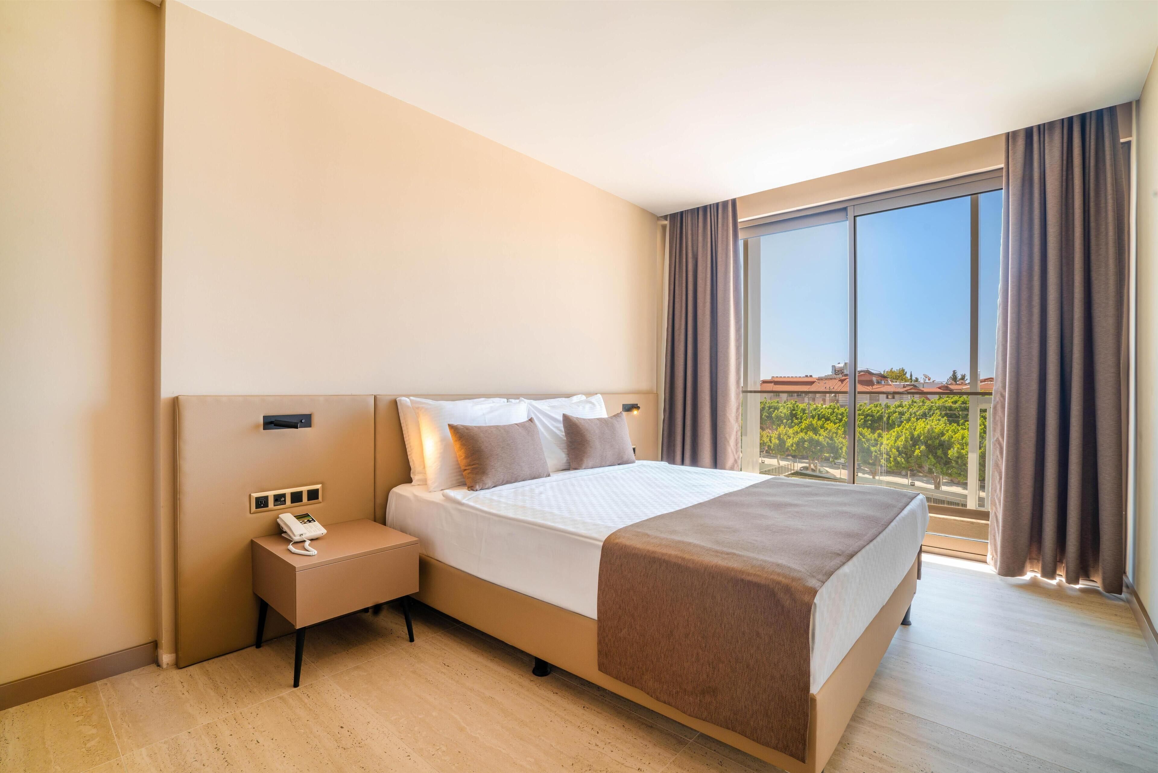 Habitación de hotel moderna con una cama grande, mesita de noche con teléfono y vistas a la ciudad a través de grandes ventanales.