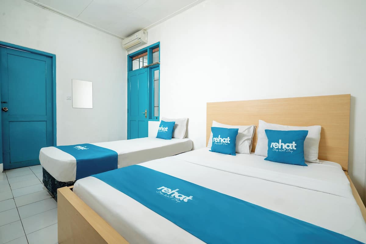 A hotel room with two single beds, each with a blue bedspread and matching pillows, and a larger bed with a wooden headboard.
