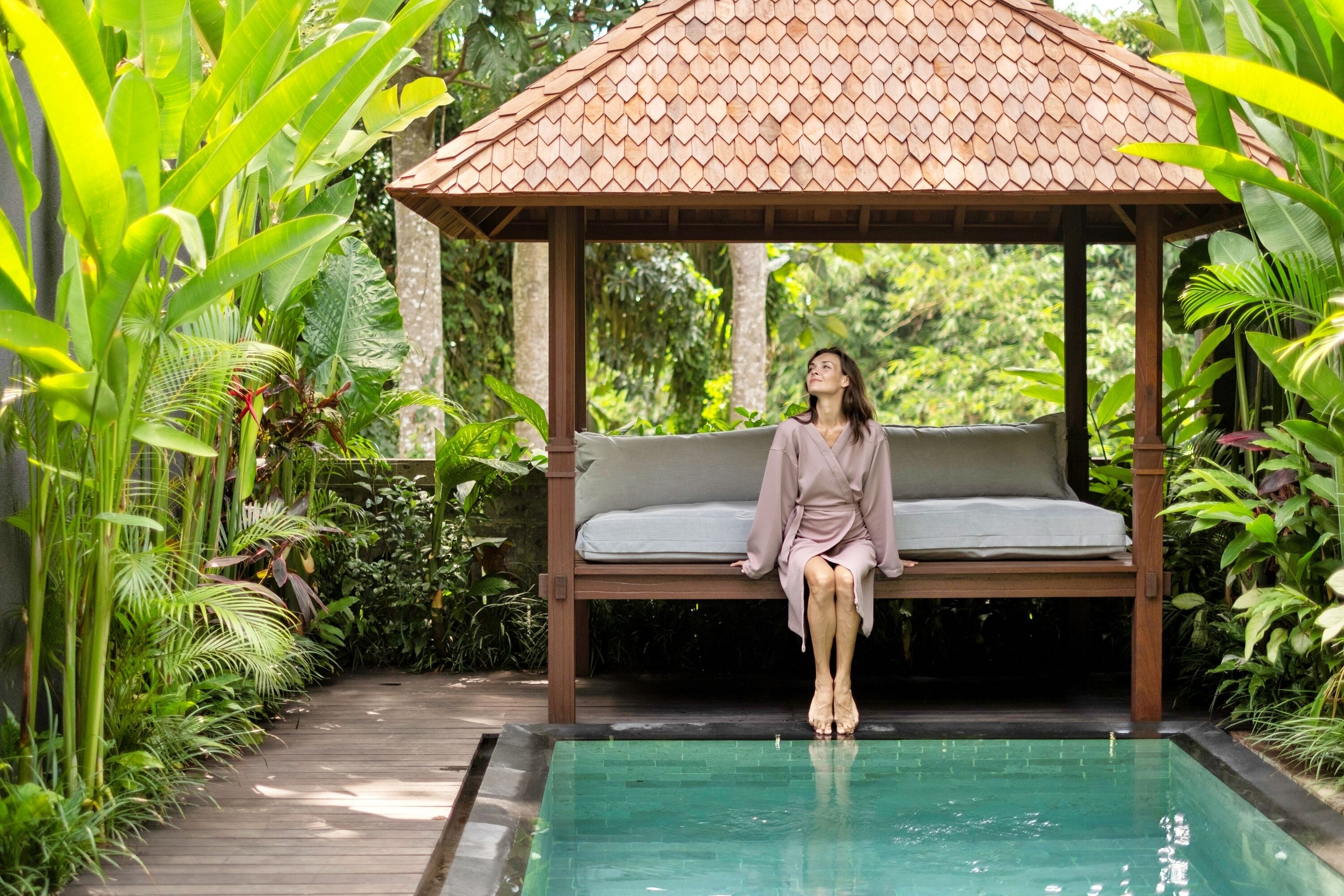 A wooden gazebo with a bench, surrounded by lush greenery and a clear pool.