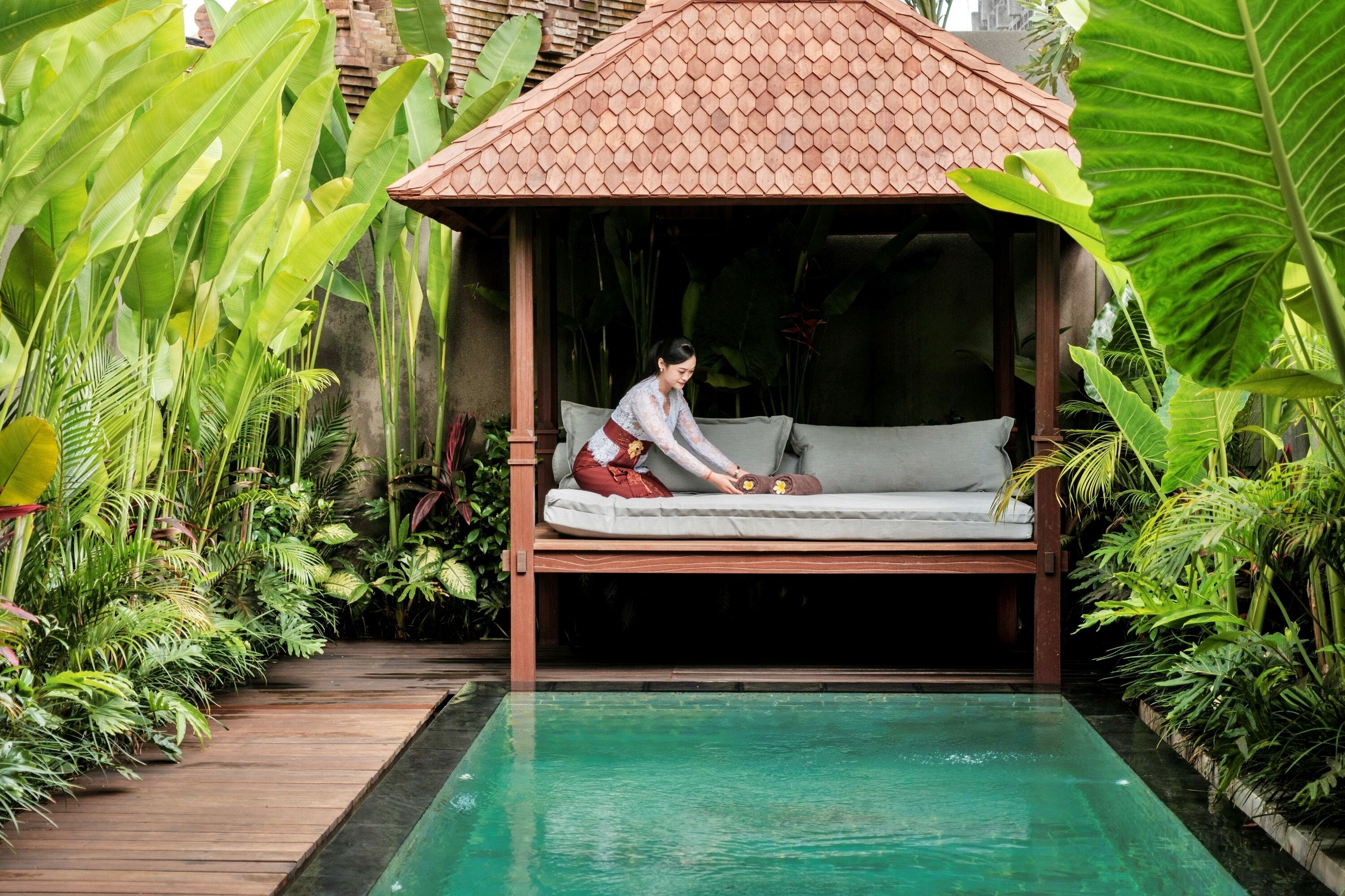 A wooden gazebo with a cushioned bench, surrounded by lush tropical plants and a clear pool.