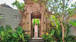 A woman in a pink dress standing at the entrance of a traditional brick structure with steps leading up to it, surrounded by lush tropical plants. - Mayaloka Villas Ubud (Ubud)