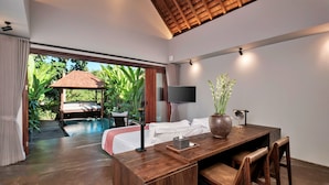 A modern room with a wooden desk, a bed with white linens, a TV mounted on the wall, and a view of a pool and garden. - Mayaloka Villas Ubud (Ubud)