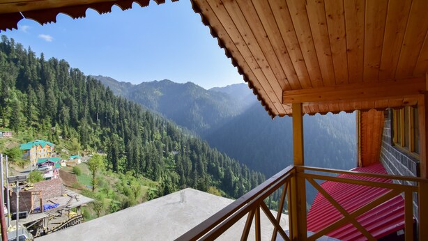 Un balcón de madera con piso rojo, que da a un paisaje montañoso con bosques densos y un pequeño pueblo abajo.