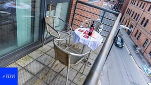 Un balcon avec un fauteuil en osier, une petite table avec une bouteille et des verres, et une vue sur une rue de la ville.