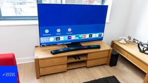 Un salon moderne avec un grand téléviseur à écran plat posé sur un meuble TV en bois, un fauteuil rouge et un petit bureau en bois agrémenté d’un téléphone et d’objets décoratifs.