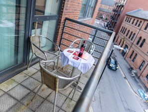 A balcony with a wicker chair, a small table with a bottle and glasses, and a view of a city street. - 2ndHomeStays I Birmingham | Ensuite | Parking&WiFi (West Midlands)
