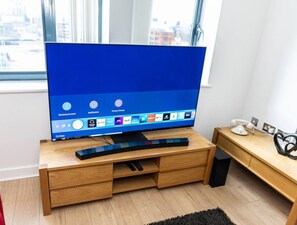 A modern living room with a large flat-screen TV on a wooden TV stand, a red chair, and a small wooden desk with a telephone and decorative items. - 2ndHomeStays I Birmingham | Ensuite | Parking&WiFi (West Midlands)