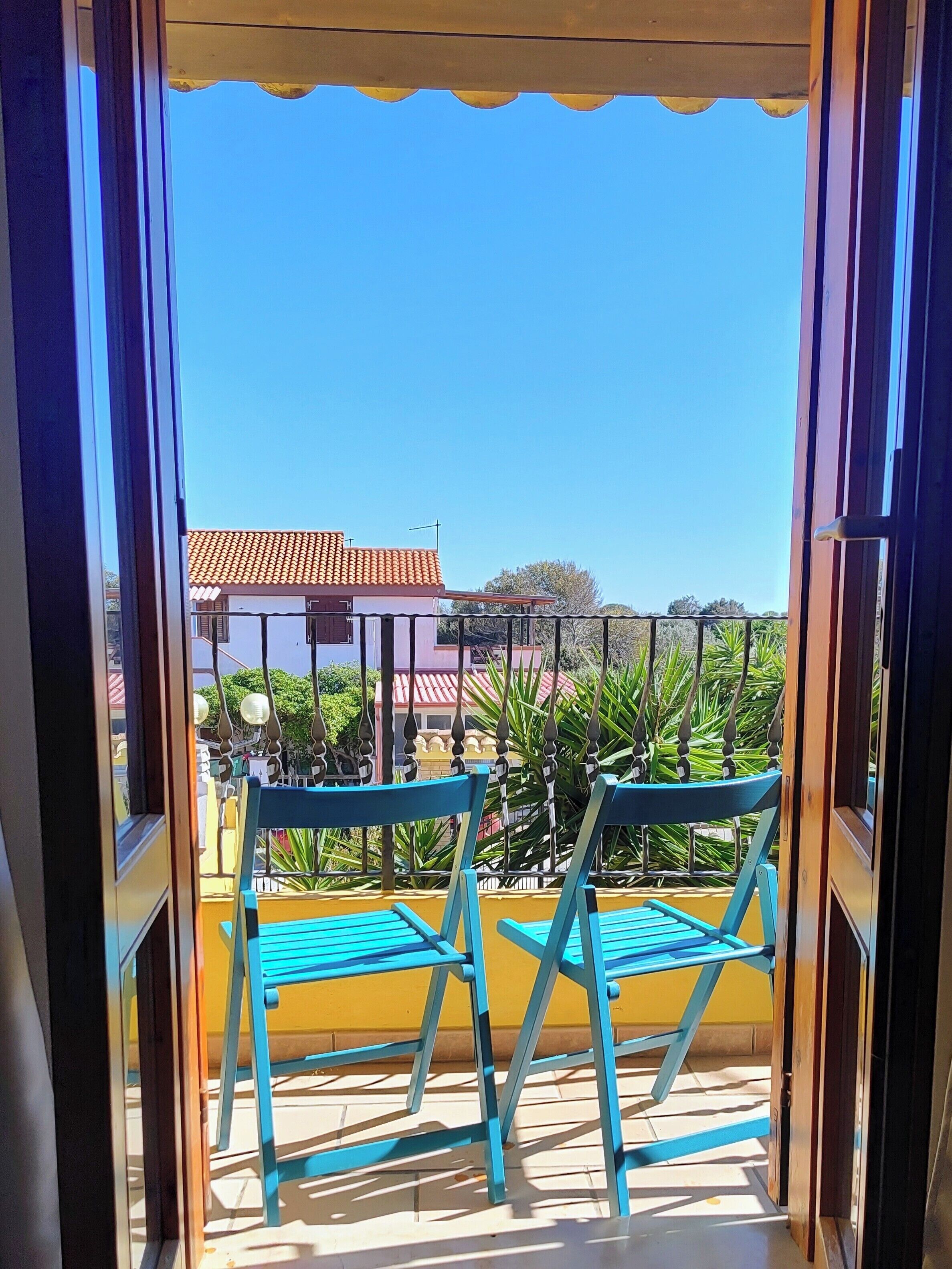Ein Balkon mit zwei blauen Stühlen, einer gelben Wand und Blick auf Gebäude mit roten Dächern.