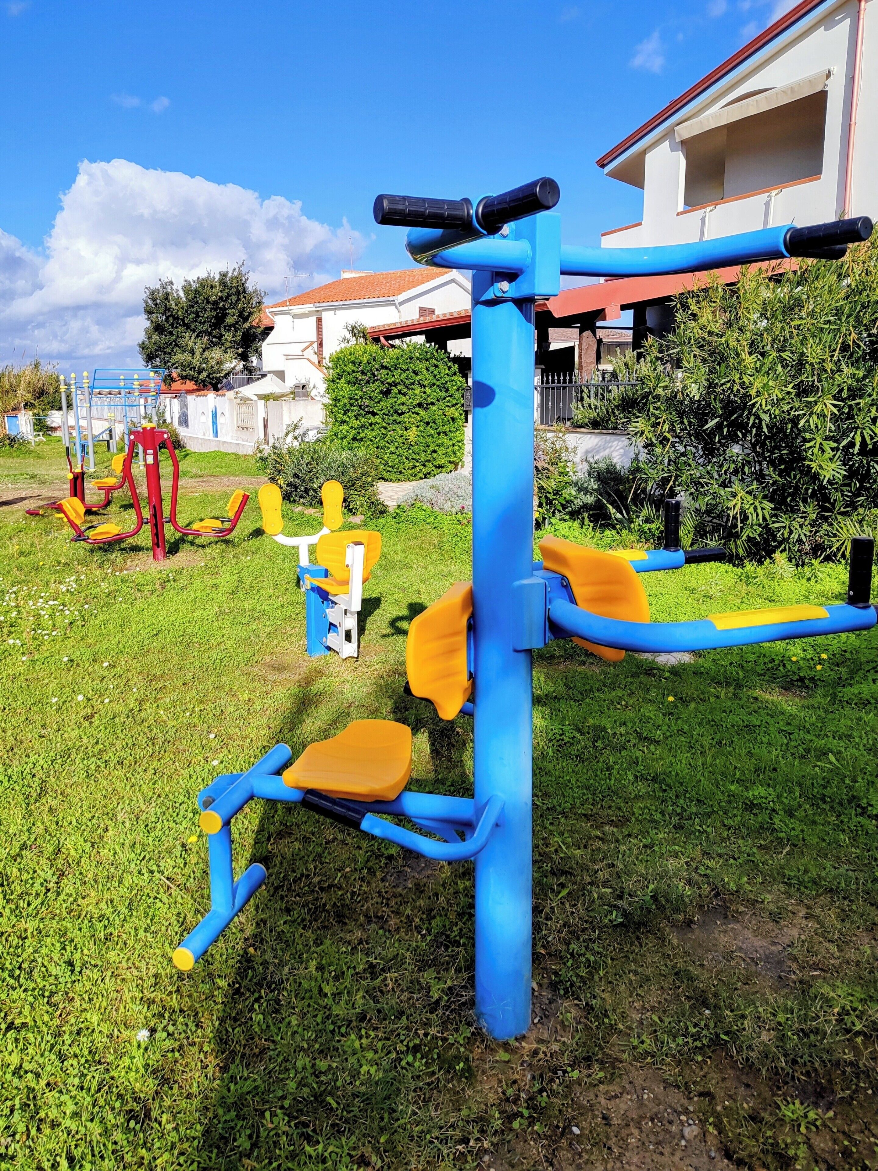 An outdoor exercise area with various fitness equipment, including a blue and yellow machine, and a red and yellow machine in the background.