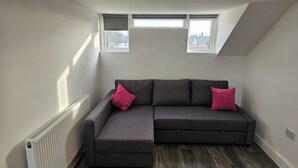 Ein Zimmer mit einem grauen Ecksofa, zwei Fenstern und einer Heizung.