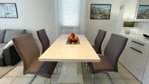 A modern dining area with a white table and four chairs, a window with blinds, and a painting on the wall. - Apartment Zlata (Jadranovo)