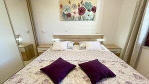 A bedroom with a floral bedspread, two purple pillows, a wooden headboard, and a painting of flowers on the wall. - Apartment Zlata (Jadranovo)