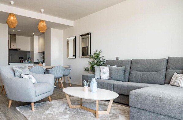 A modern living room with a grey sofa, a matching armchair, a coffee table, and a dining area with chairs and a table.