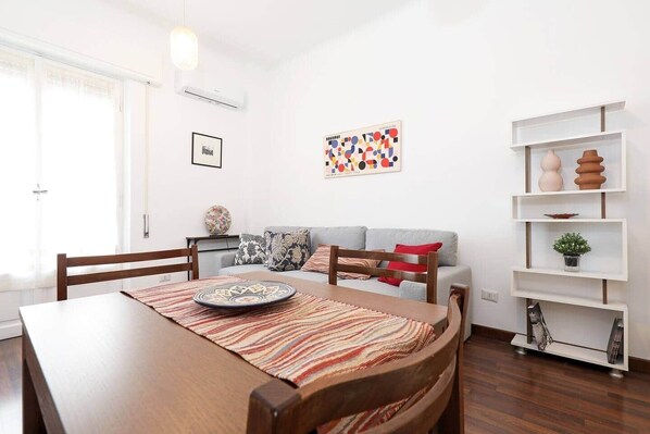 A dining area with a wooden table, chairs, a patterned rug, a grey sofa with cushions, a wall-mounted shelf with decorative items, and a framed picture.