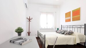 A bedroom with a bed, a bedside table with a book and a plant, a coat rack, and a window with curtains.
