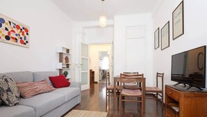 A living room with a grey sofa, wooden chairs, a TV, and a painting on the wall.