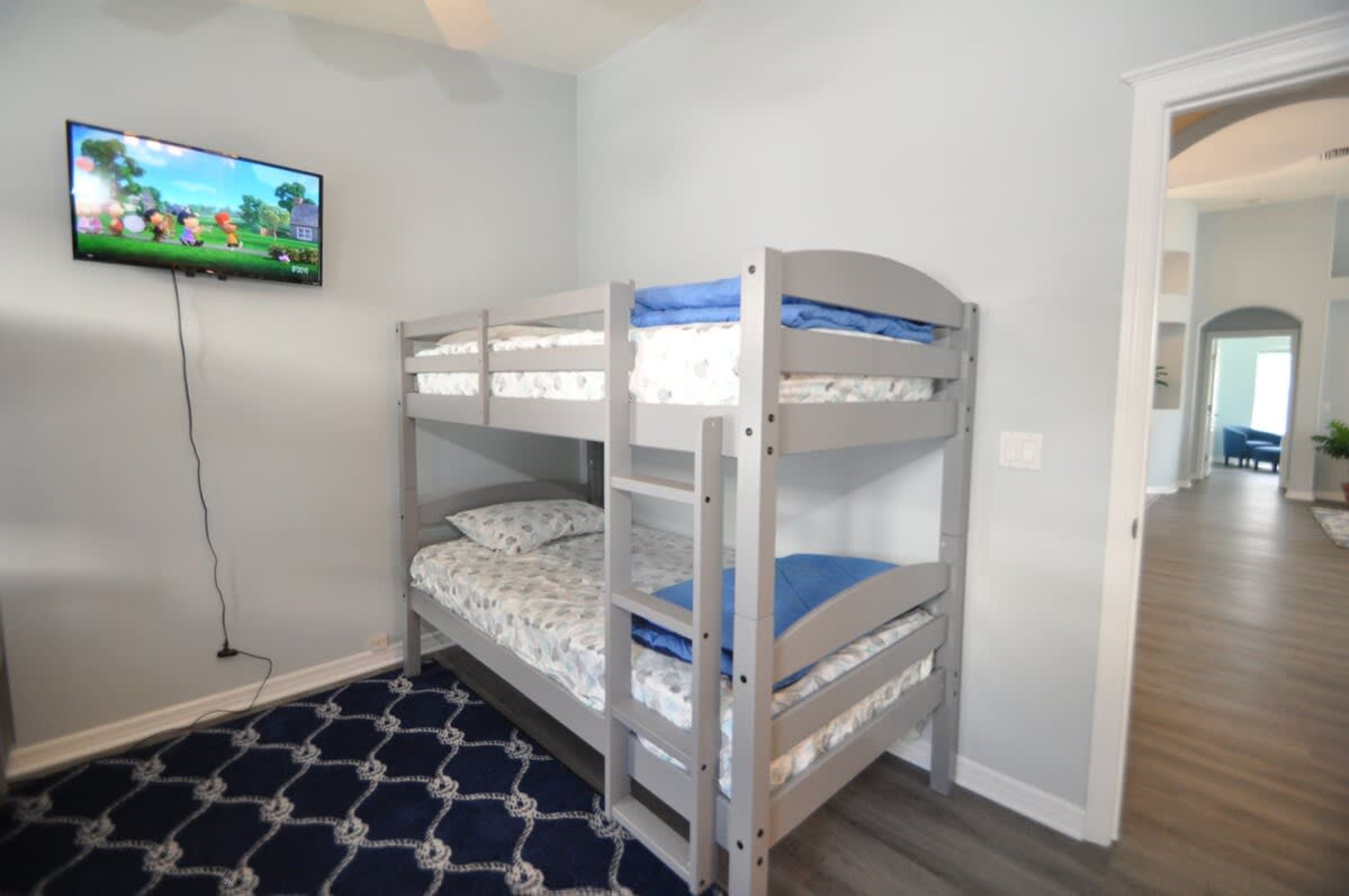 Quarto com beliche, televisão na parede, tapete estampado e corredor que dá para outro quarto.