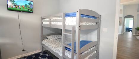 Quarto com beliche, televisão na parede, tapete estampado e corredor que dá para outro quarto.