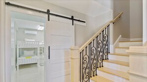 Un escalier avec une rampe en fer forgé noir, des murs blancs et une porte blanche donnant sur une pièce où se trouve un lit superposé.
