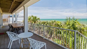 Un espacio al aire libre techado con una mesa de madera y sillas blancas, con vistas a una playa con palmeras y agua azul cristalina.