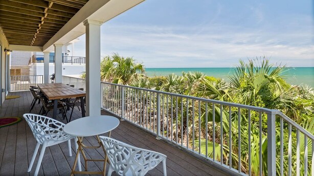 Un espace extérieur abrité, doté d’une table en bois et de chaises blanches, qui offre une vue sur une plage bordée de palmiers et d’une eau d’un bleu cristallin.