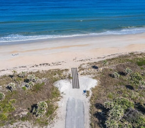 An aerial view of a beach with a paved path leading to the water, surrounded by sparse vegetation and a clear sky.