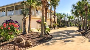 Un hotel con una galería techada, palmeras y macizos de flores.