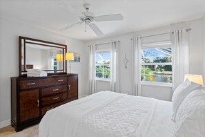 Une chambre à coucher comprenant un lit, une commode, un ventilateur de plafond et deux fenêtres avec des rideaux.