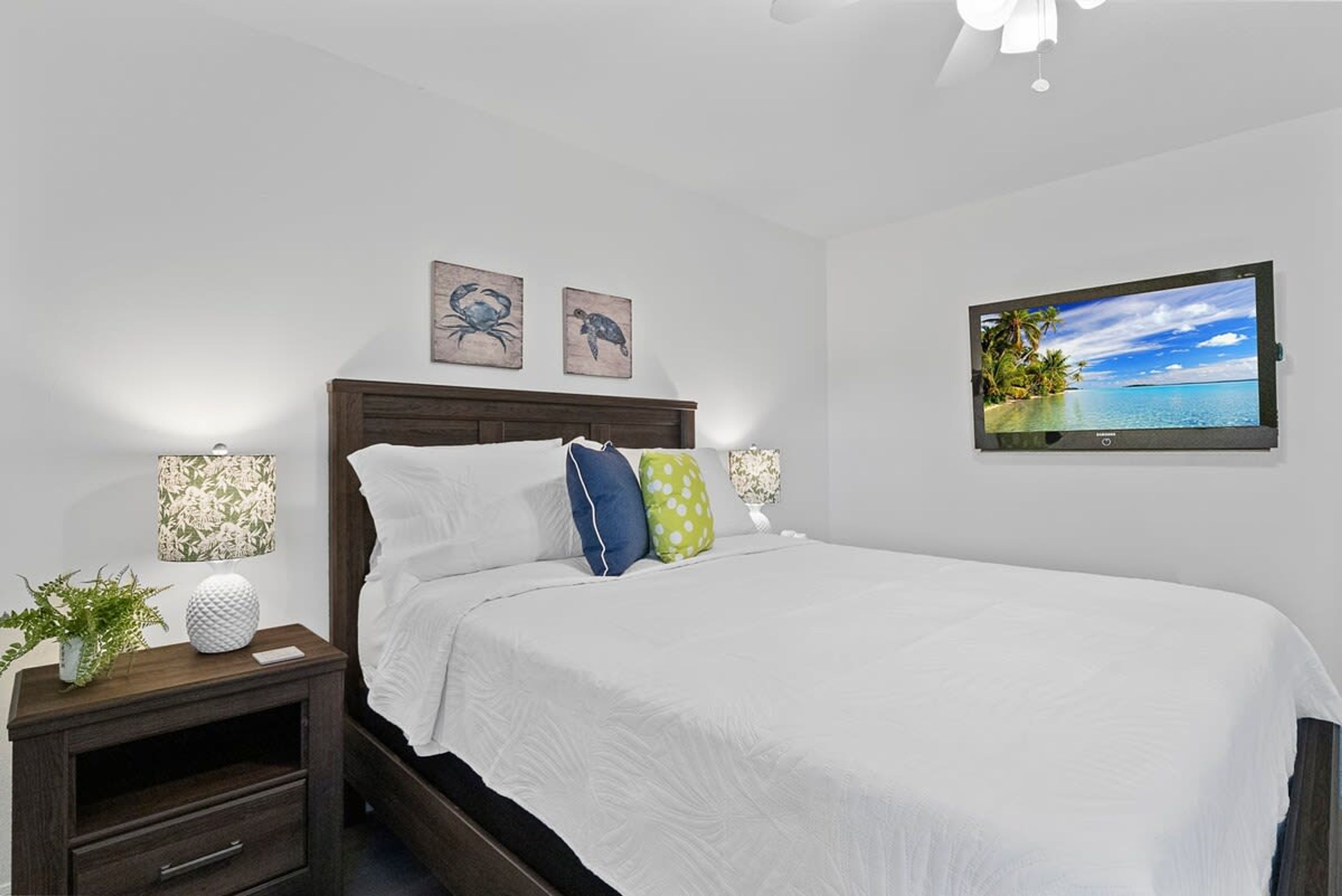 A neatly made bed with white bedding and decorative pillows, a wooden nightstand with a lamp and a plant, and a wall-mounted TV displaying a tropical beach scene.