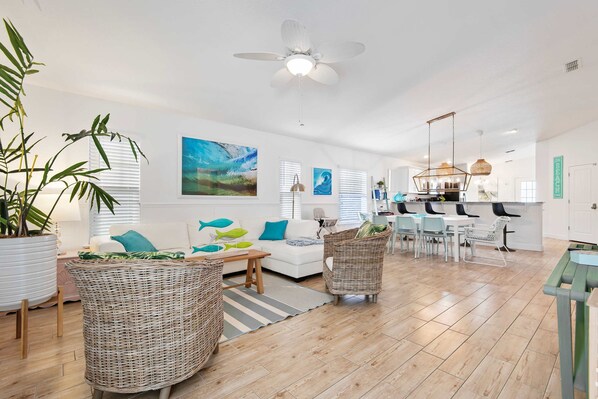 A spacious living room with a white sofa, wicker chairs, and a dining area with a kitchen in the background.