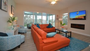 A modern living room with a large orange sectional sofa, a blue patterned armchair, and a television mounted on the wall. - Beach Front 5 bed 4 Bath Luxury House Hot Tub Fenced Yard Pets Welcome (Palm Coast)