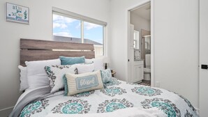 Een slaapkamer met een houten hoofdbord, een groot raam, een bed met beddengoed met een patroon en een badkamer die door een open deur zichtbaar is.