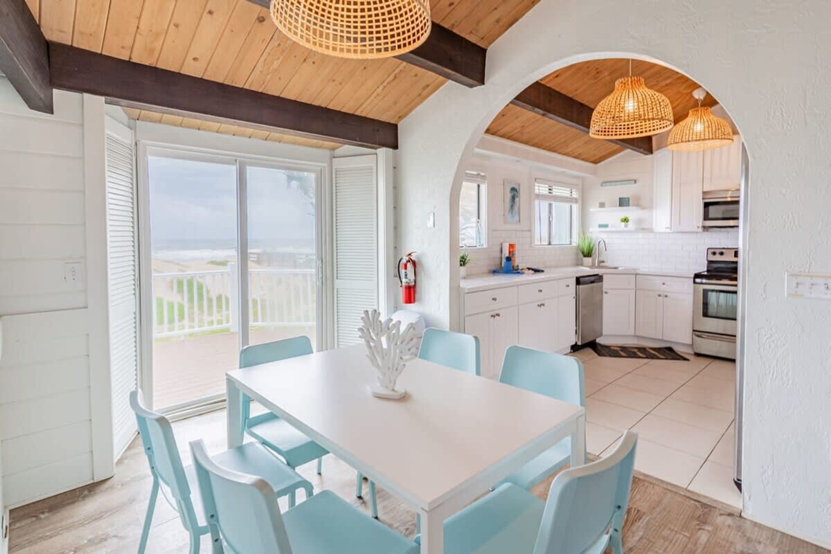A modern kitchen with white cabinets, a white countertop, and a built-in oven. There is a dining area with a white table and blue chairs, and a large window overlooking the beach.