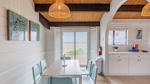 A dining area with a white table and light blue chairs, a view of the beach through open shutters, and a kitchen with white cabinets and a window. - Pelican Beach Private Ocean Front Beach House- Sleeps 10 Bring Your Surf Boards (Palm Coast)