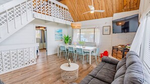 A spacious living area with a wooden ceiling, a gray sofa, a dining table with chairs, and a staircase with a white railing. - Pelican Beach Private Ocean Front Beach House- Sleeps 10 Bring Your Surf Boards (Palm Coast)