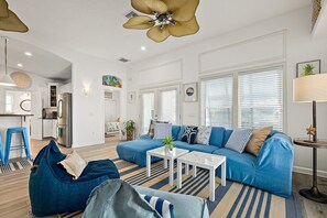 Ein modernes Wohnzimmer mit einem blauen Sofa, einem weißen Couchtisch und blauen Sitzsäcken. Im Hintergrund befindet sich eine Küche mit weißen Schränken und Edelstahlgeräten.