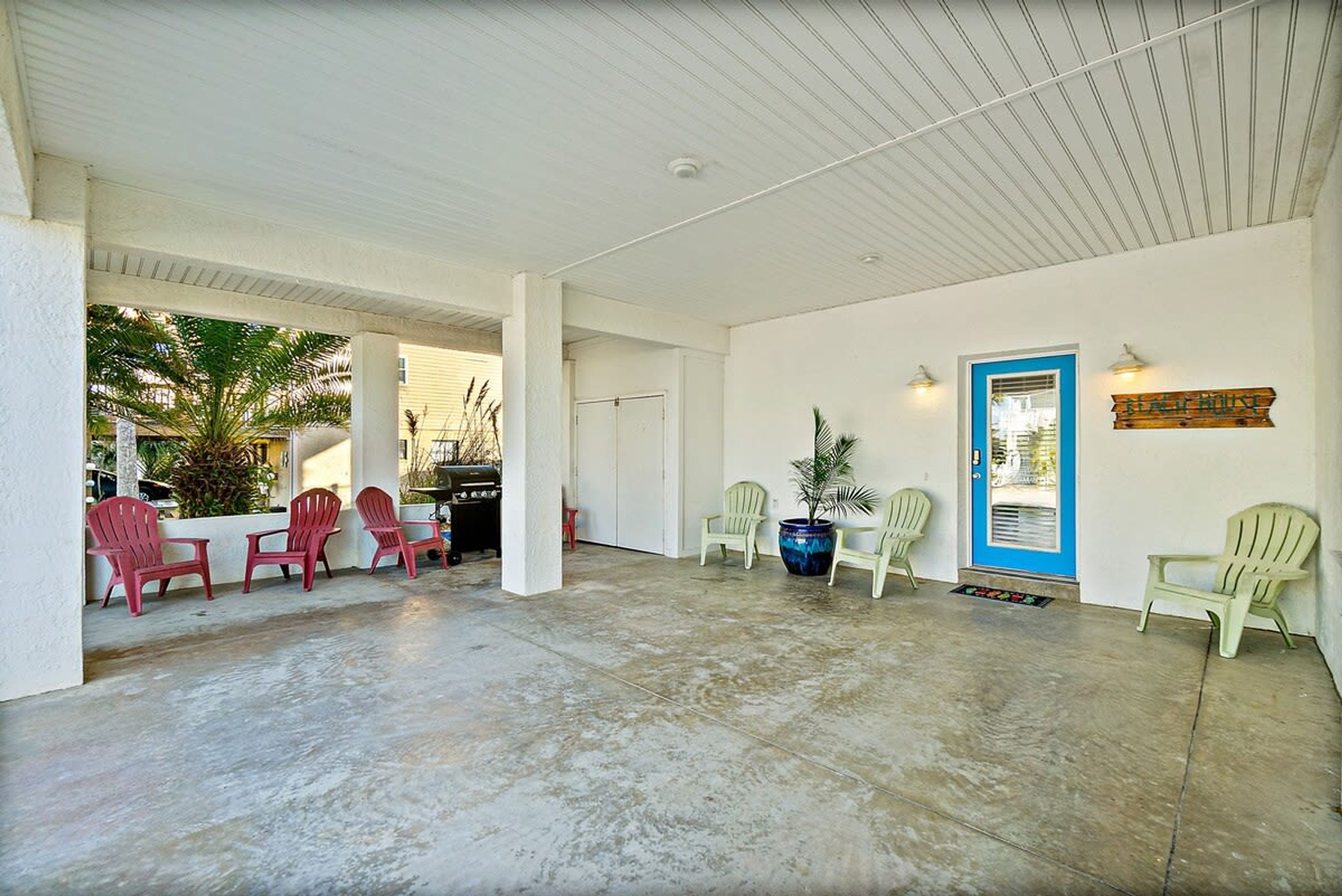 A covered outdoor area with concrete flooring, white walls, and a blue door. There are several plastic chairs and a potted plant.
