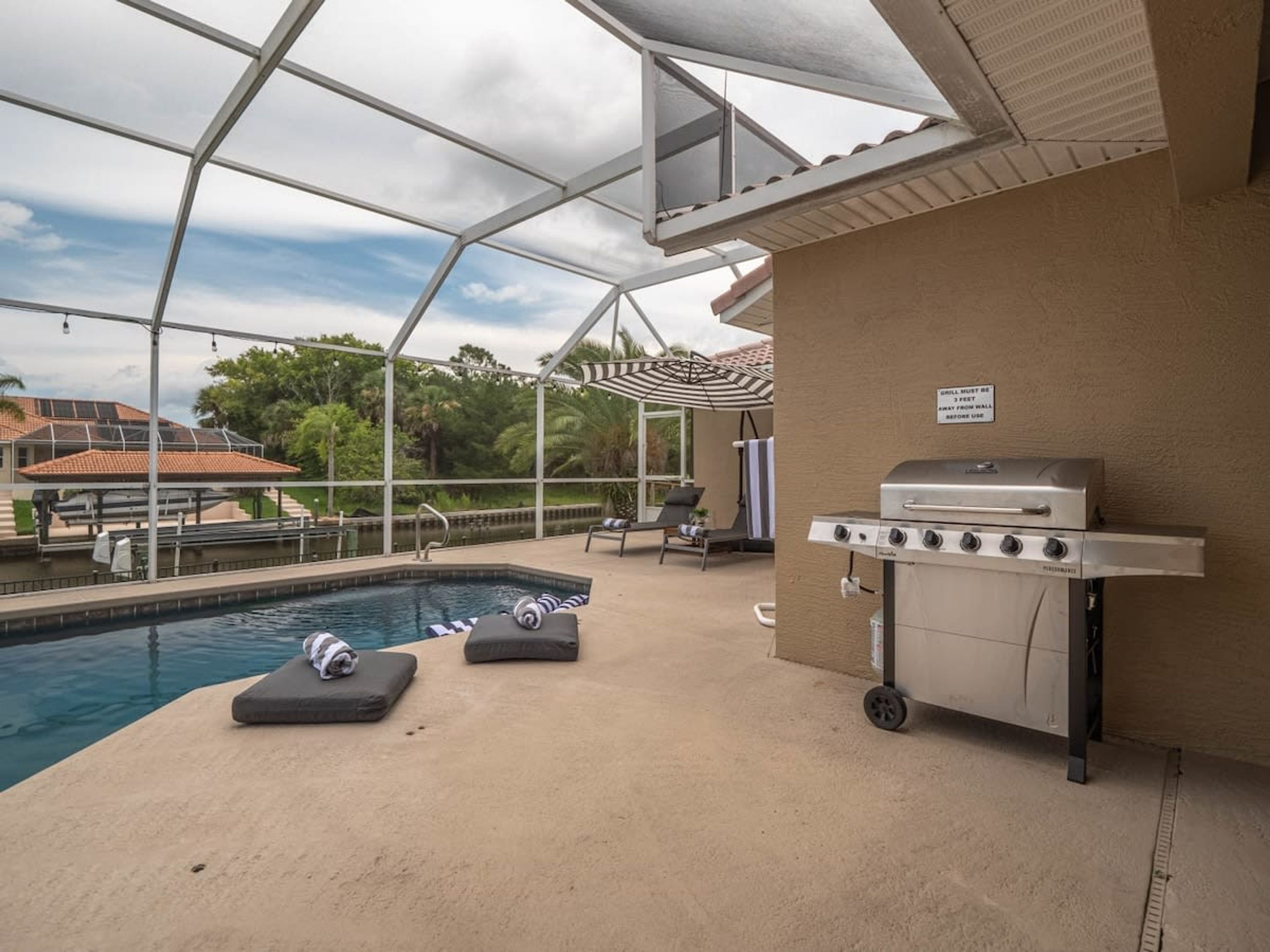 An outdoor pool area with a covered patio, lounge chairs, and a grill.