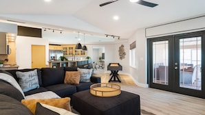 A modern living room with a sofa, ottoman, and a pool table. There is a ceiling fan and recessed lighting.