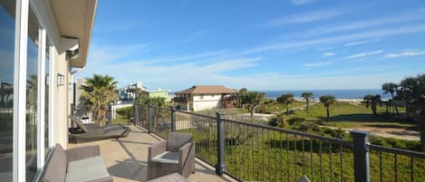A balcony with wicker furniture, a view of the ocean, and a grassy area with palm trees.
