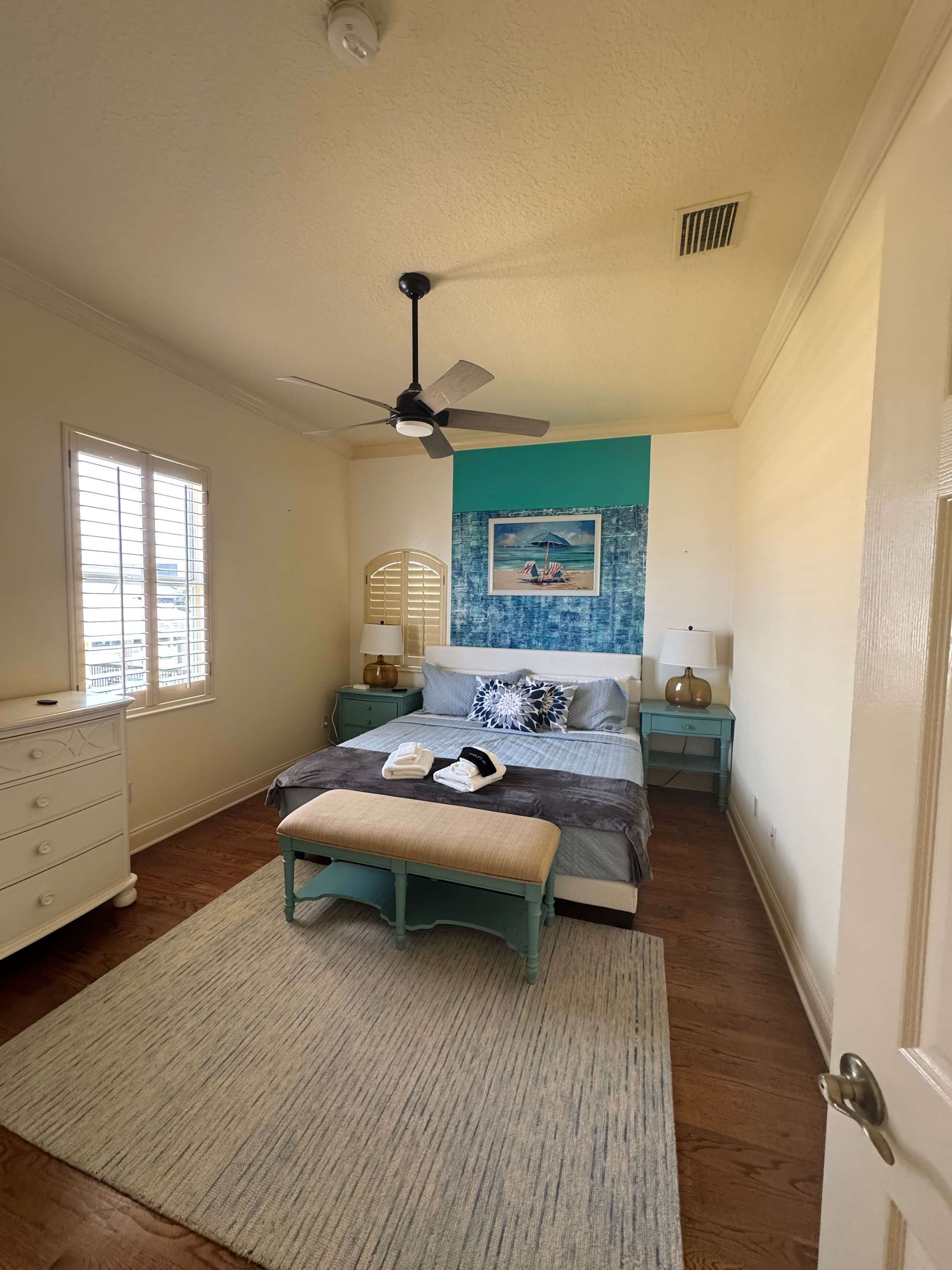 A bedroom with a bed, a bench, a dresser, a ceiling fan, and a window with blinds.